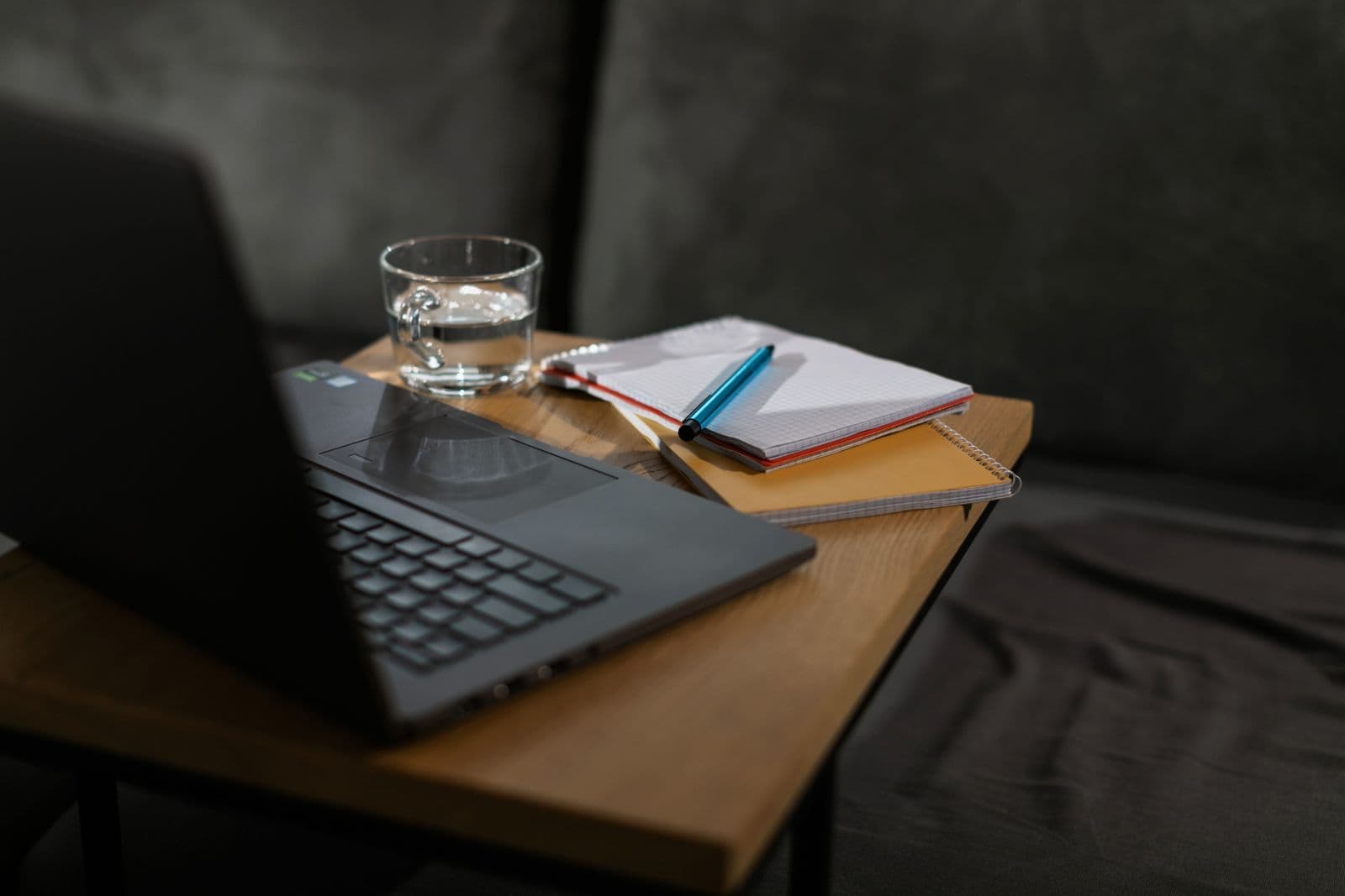 An open laptop next to several notebooks and a glass of water on a wooden table