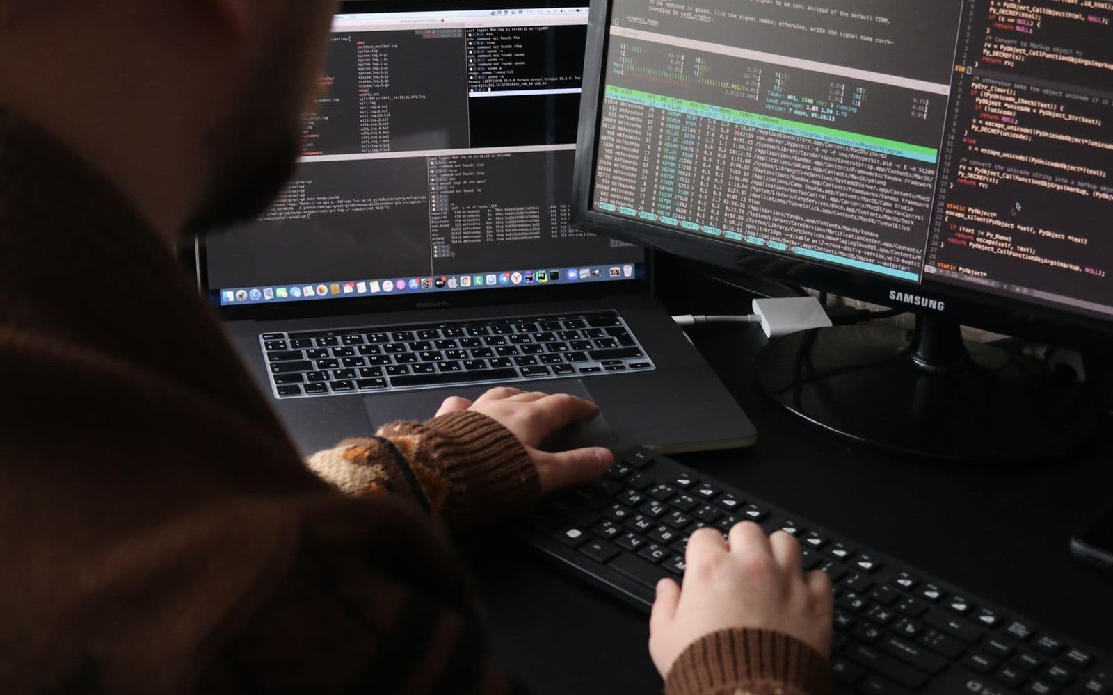 Person tippt an einem Arbeitsplatz mit zwei Monitoren und einem Laptop, auf allen Bildschirmen steht Code