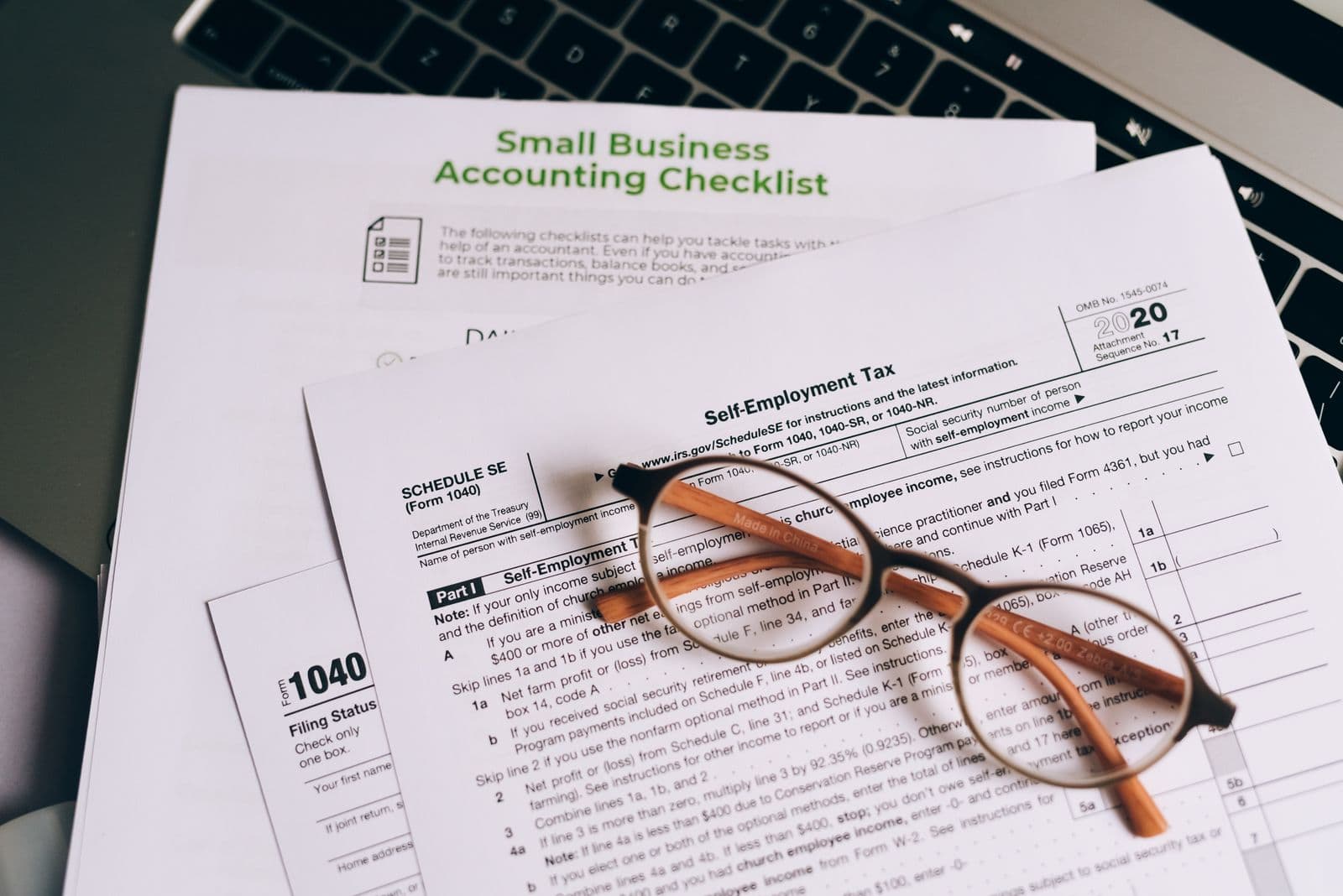 Tax forms and a small business accounting checklist next to a laptop, with glasses resting on top