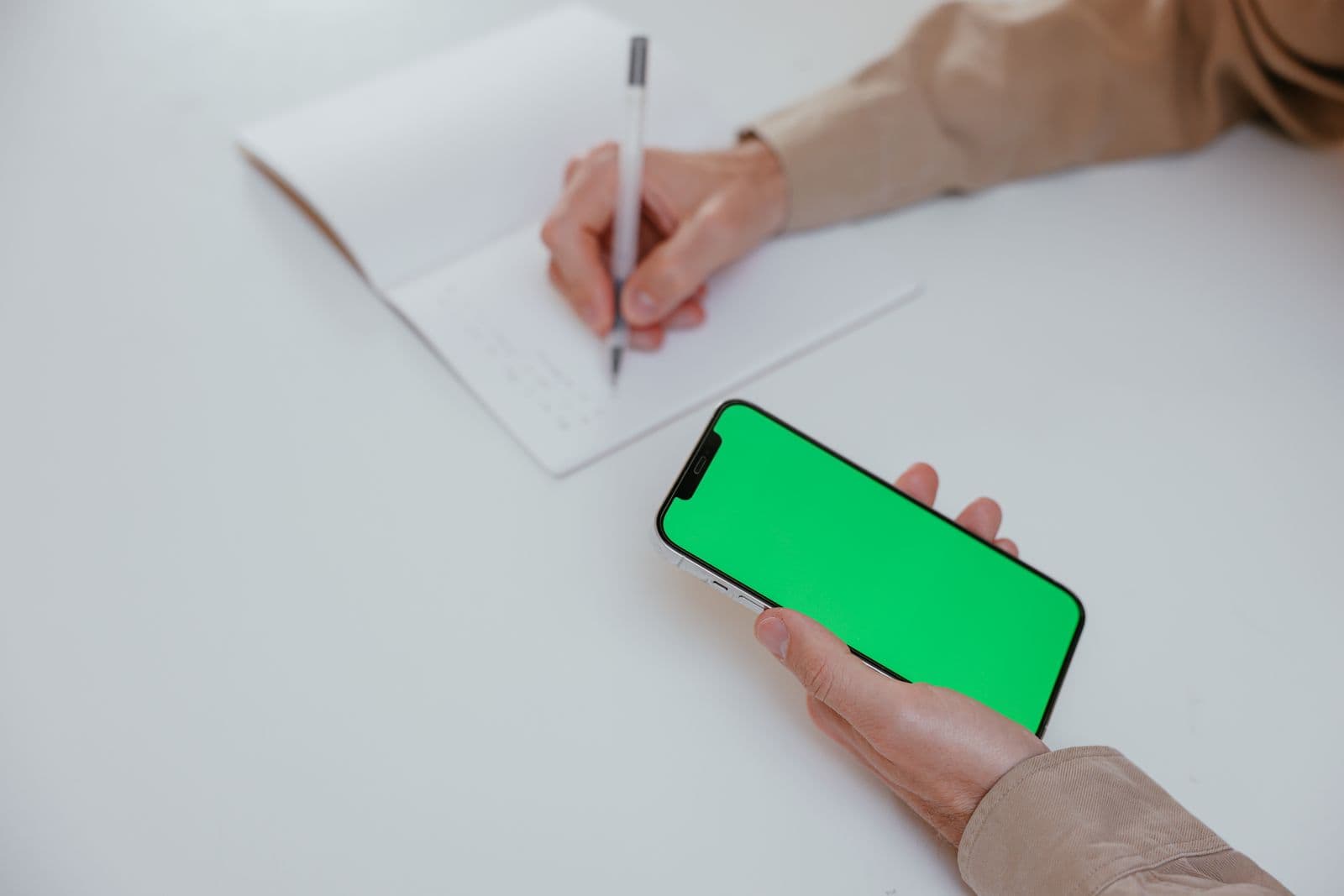 Close-up of a hand writing notes with a pen on a notepad while the other hand holds a smartphone