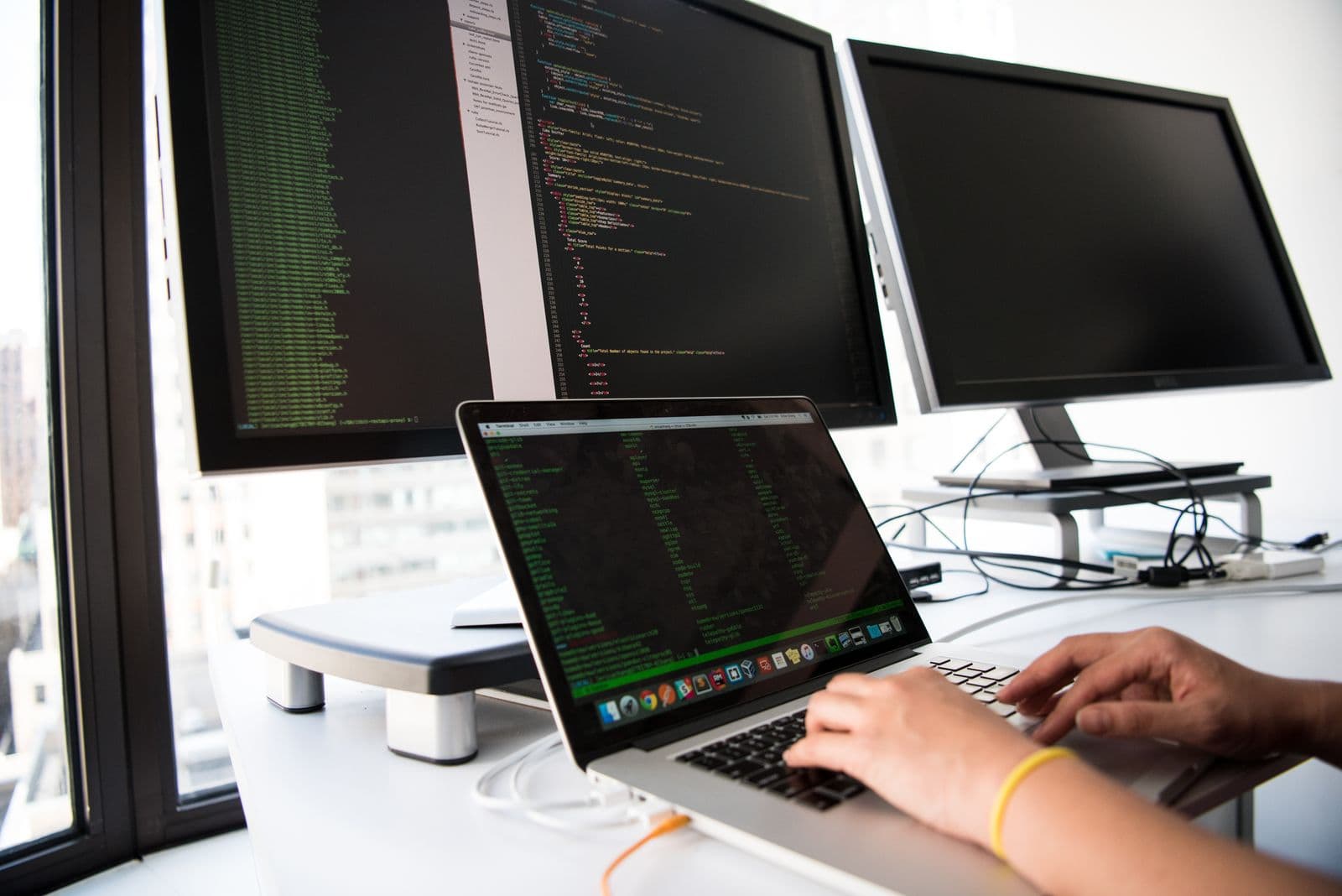 A developer types on a laptop with multiple code-filled monitors in the background in an office