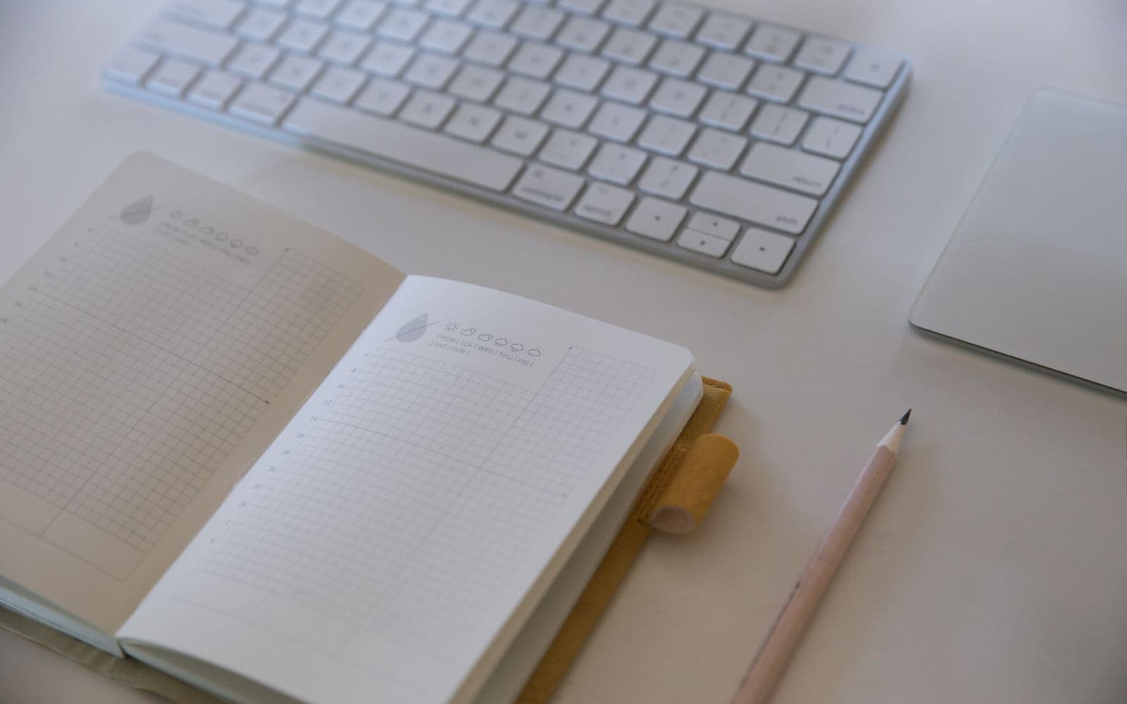 Aufgeschlagenes punktkariertes Notizbuch neben einer weißen Tastatur und einem Bleistift auf einem hellen Schreibtisch