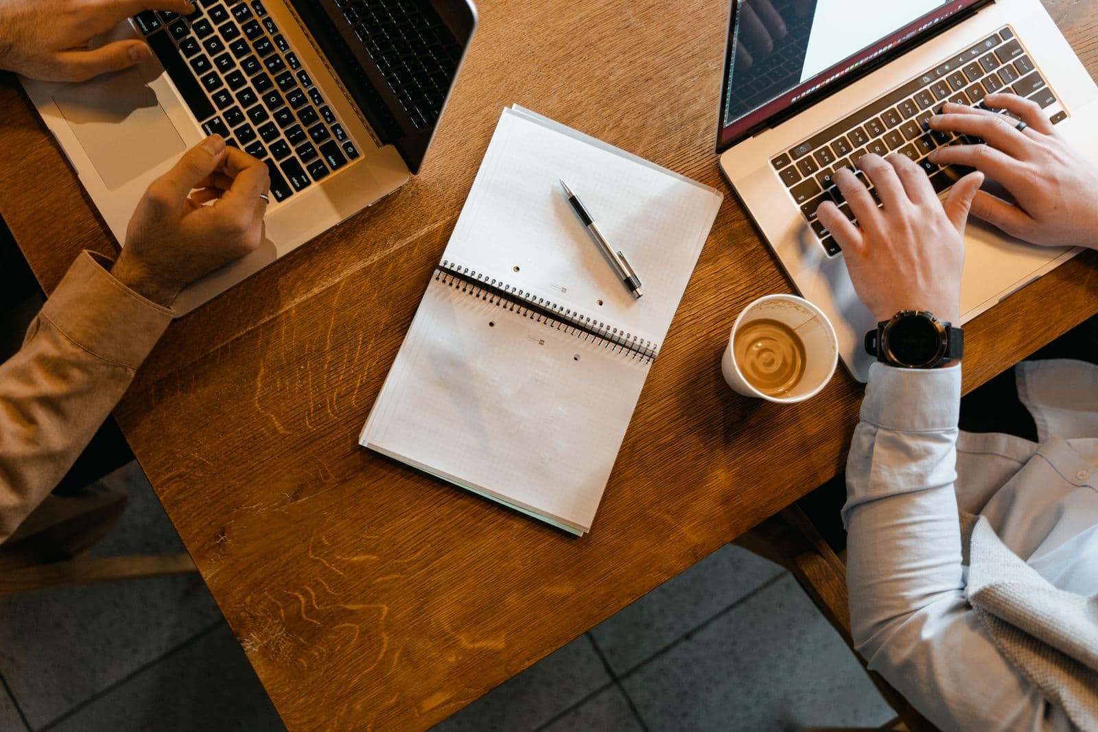 Draufsicht auf einen Holzschreibtisch mit zwei Laptops, Notizblöcken und einer Kaffeetasse zwischen zwei Personen
