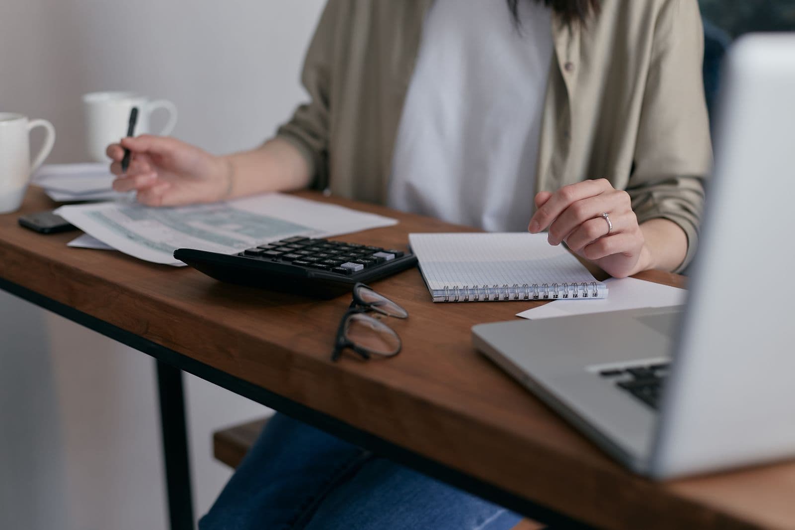 Eine Frau erledigt an einem Holzschreibtisch die Finanzen mit Taschenrechner, Belegen, zwei Kaffeetassen und einem Laptop