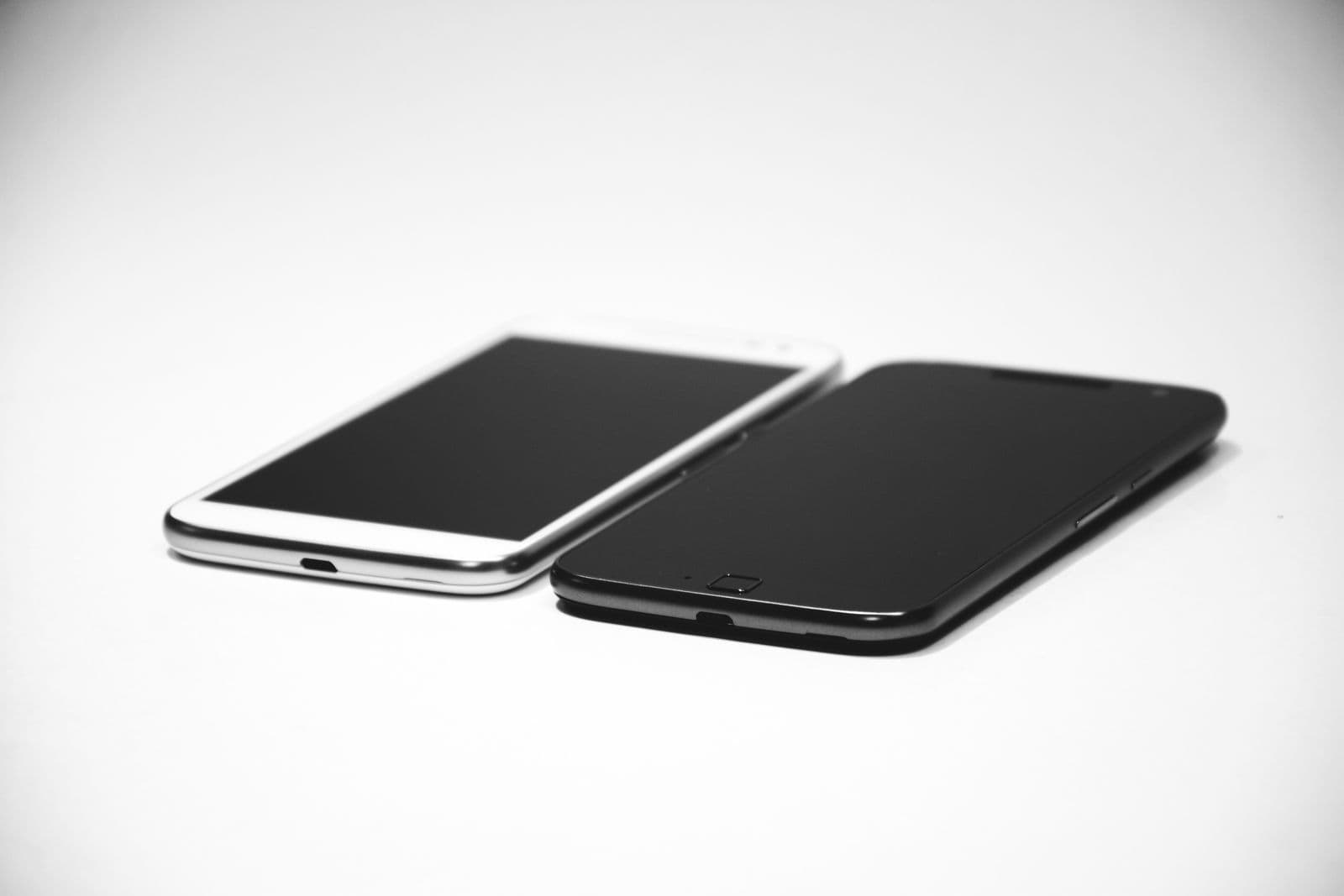 Zwei Smartphones, eines schwarz und eines weiß, mit ausgeschaltetem Display nebeneinander auf weißem Untergrund