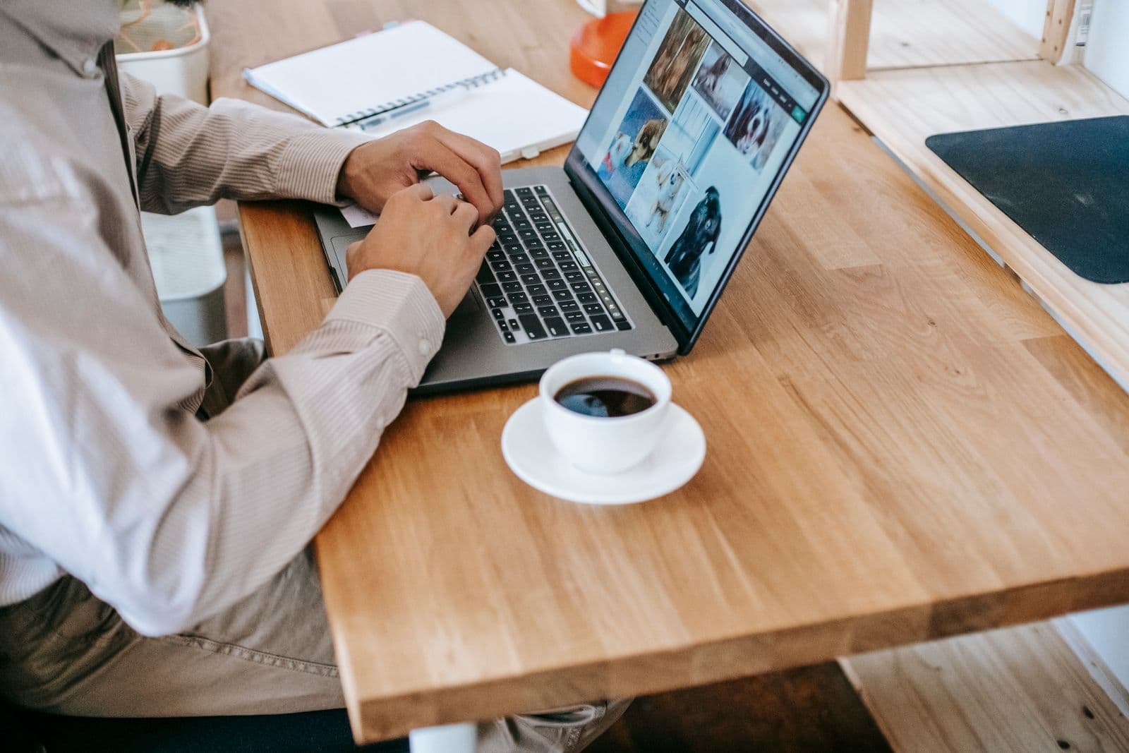 Person tippt am Laptop und trinkt Kaffee, daneben liegt ein Notizbuch auf einem Holztisch