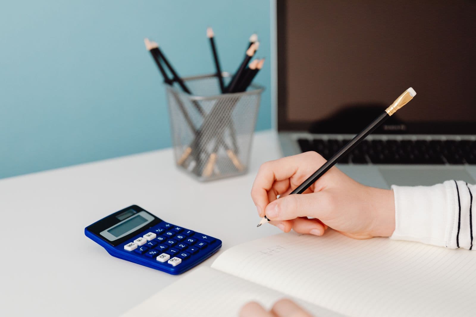 Hand writing in a notebook next to a calculator and a laptop on a desk