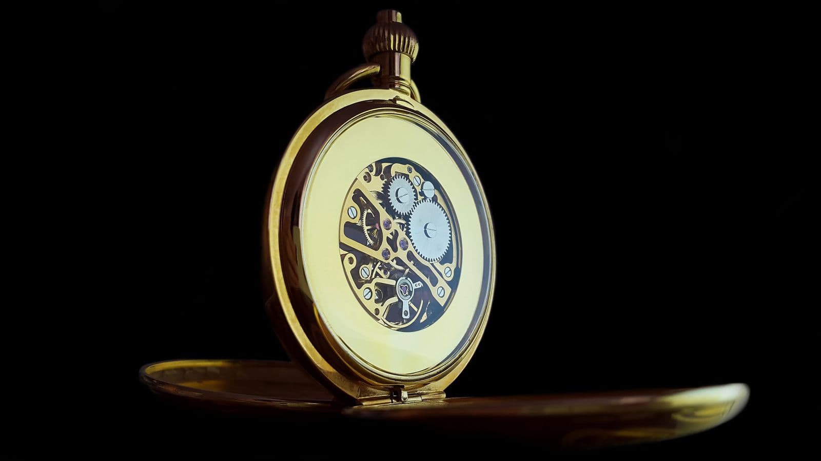 Geöffnete goldene Taschenuhr mit Blick auf das freiliegende Uhrwerk mit Zahnrädern vor schwarzem Hintergrund