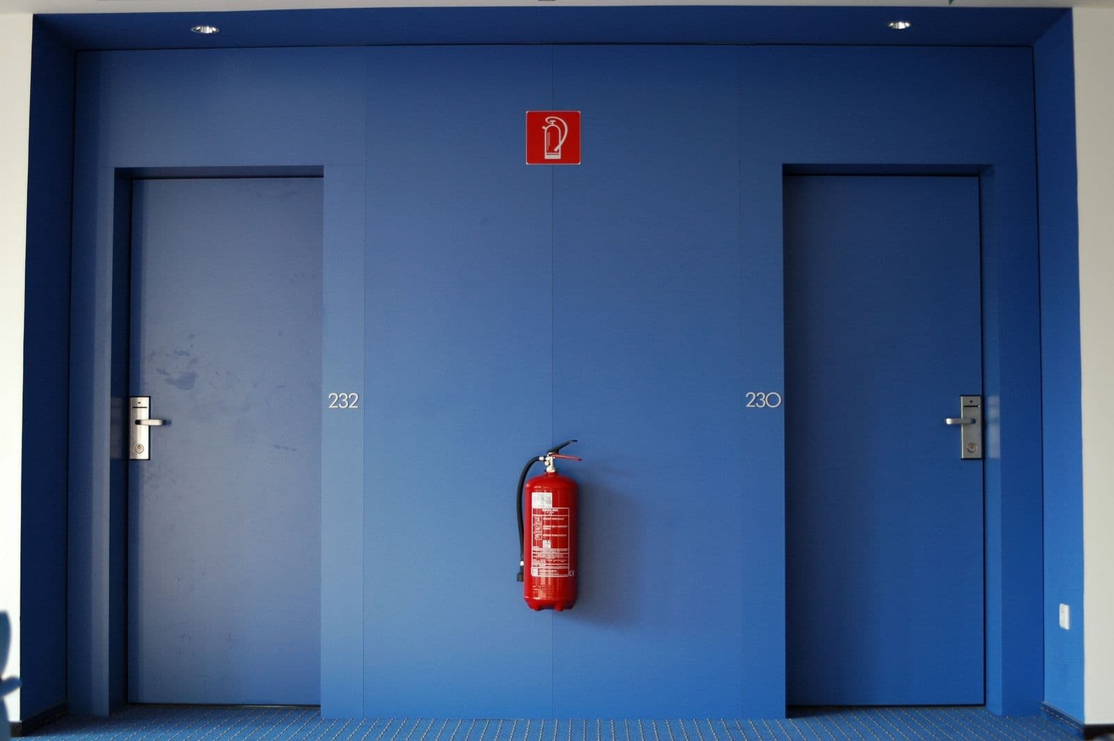 Zwei nummerierte Türen mit elektronischen Schlössern in einem blauen Flur, dazwischen ein Feuerlöscher an der Wand