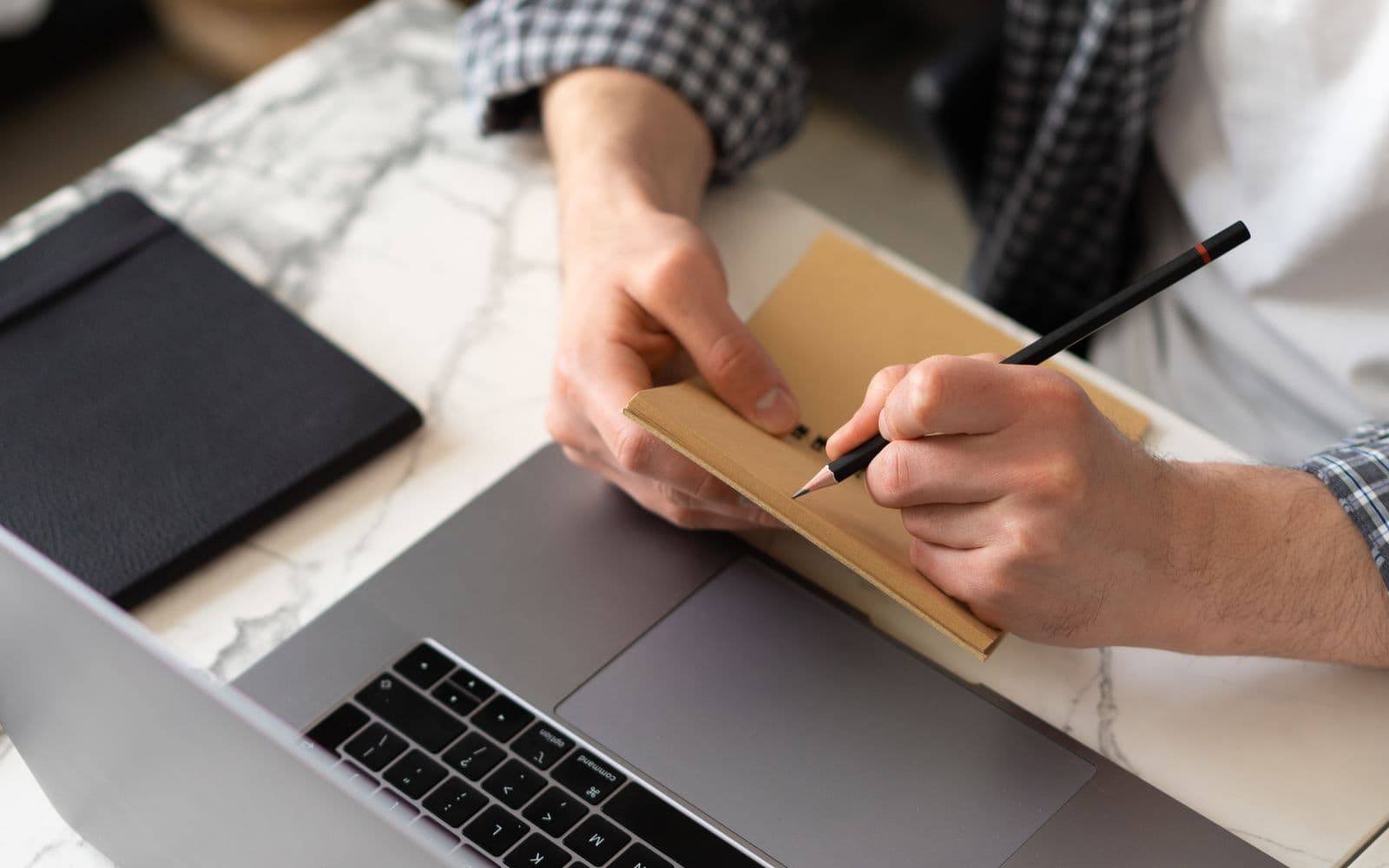 Person macht Notizen in einem Notizbuch über der Laptop-Tastatur