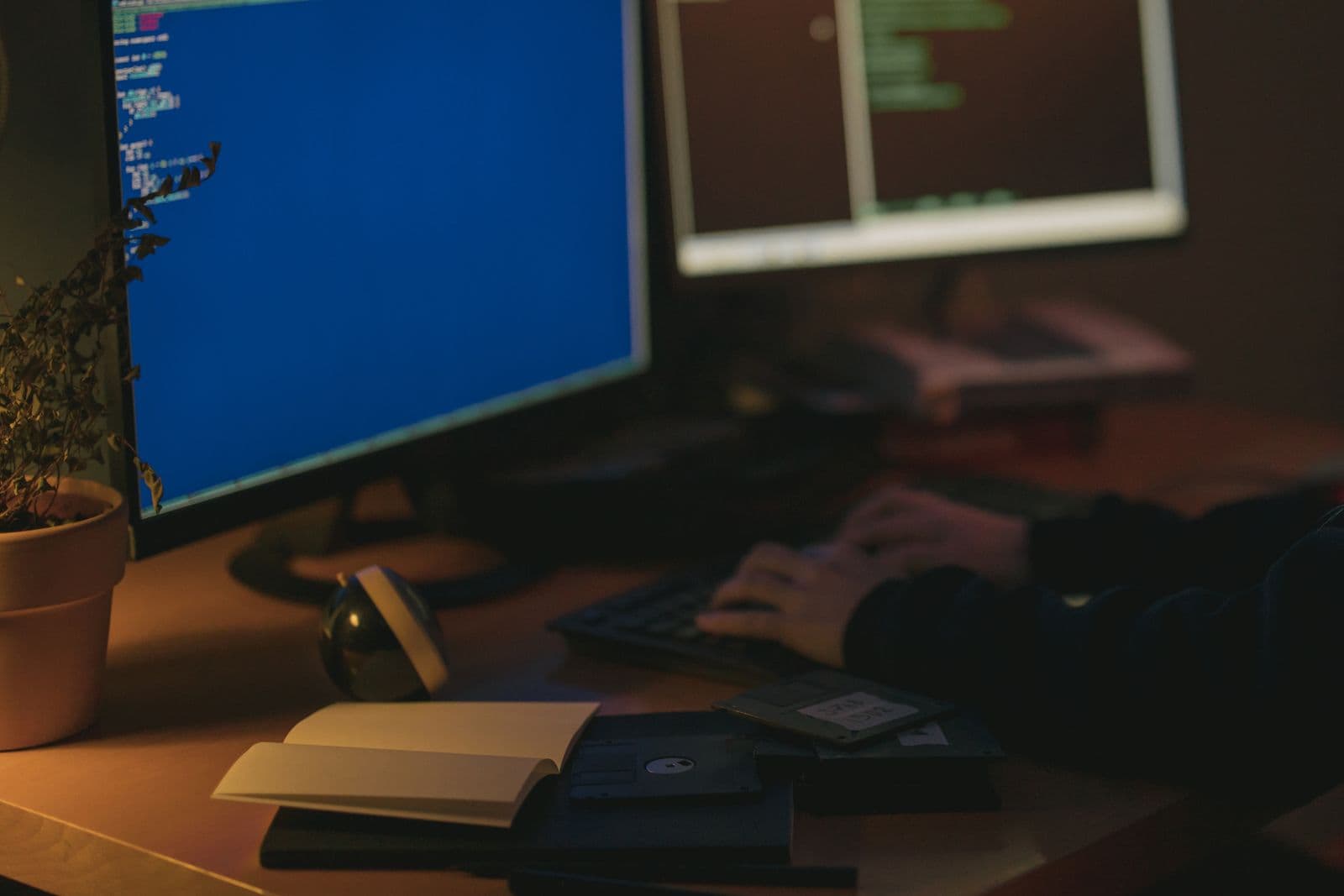Zwei Bildschirme mit Code nebeneinander auf einem Schreibtisch, im Vordergrund eine tippende Hand